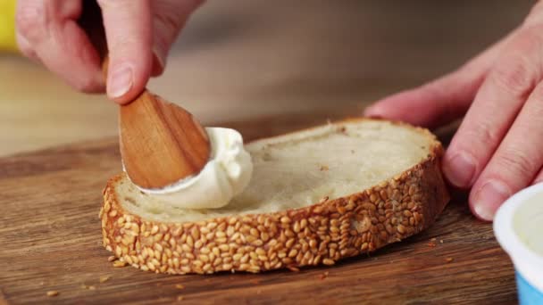 Женские руки наносят свежий сливочный сыр на ломтик свежеиспеченного хлеба крупным планом. домашняя кухня завтрак — стоковое видео