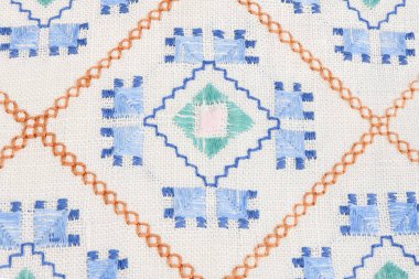 Beyaz keten üzerine geleneksel el yapımı nakışlar yakın plan. Mavi, yeşil ve turuncu iplikli Geometrik folklor süslemeleri elmas şekiller oluşturuyor. Tekstil arkaplanı.