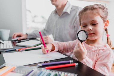 Kızı, babasıyla masaya otururken resim çiziyor. 