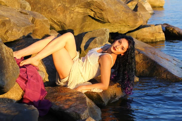 Beautiful young woman sitting on the stone on the seashore — Stock Photo, Image