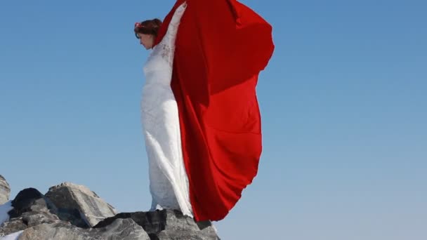 Sposa si trova sulla scogliera con un impermeabile rosso fluente sul vento contro il cielo blu e lo sfondo invernale . — Video Stock