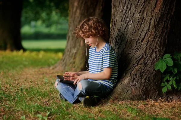 El chico con la tableta en las manos se sienta bajo un gran árbol . — Foto de Stock