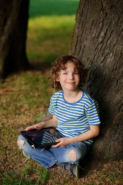 El niño de 8-9 años se sienta apoyado contra un árbol y sostiene la tablilla en la mano . — Foto de Stock