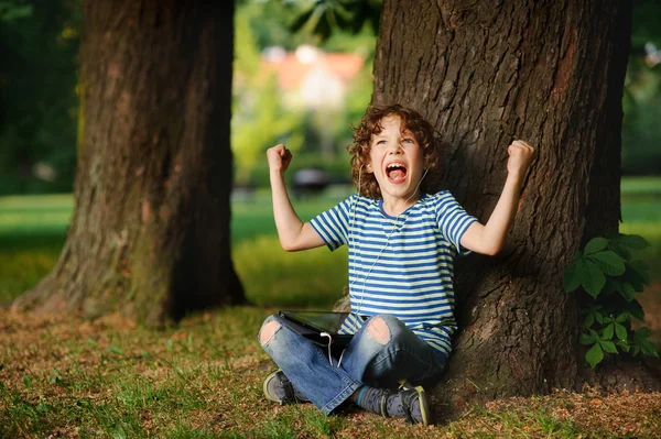 El muchacho indignado se sienta debajo de un árbol con la tableta en el regazo . — Foto de Stock