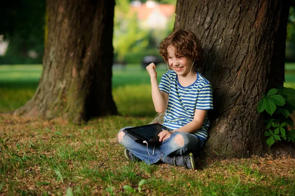 El niño de 8-9 años se sienta, apoyado contra el árbol y sostiene la tablilla en un regazo . — Foto de Stock