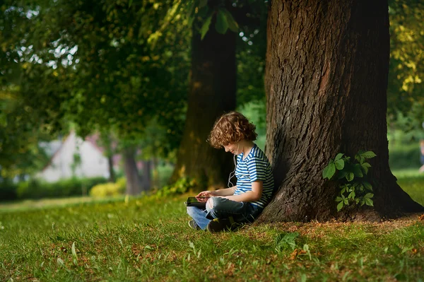 El pequeño se sienta debajo de un gran árbol y mira la pantalla de la tableta . — Foto de Stock