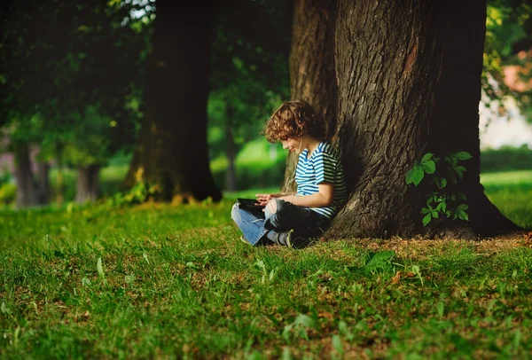The boy sits having crossed legs near a big tree.