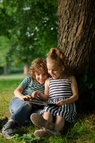 Broer met zuster van 7-9 jaar onder een boom zitten en kijken in de tablet. — Stockfoto