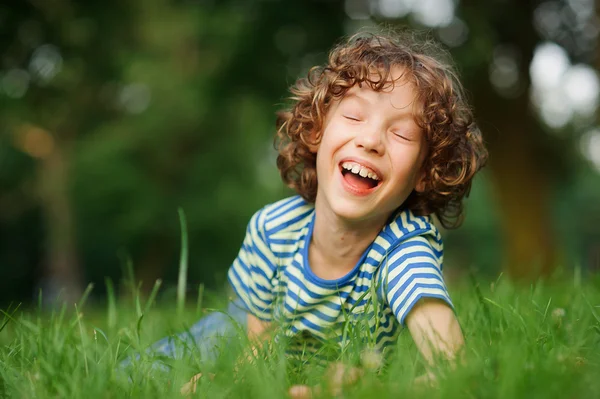 Худыш 8-9 лет лежит в зеленой траве и громко смеется. . — стоковое фото