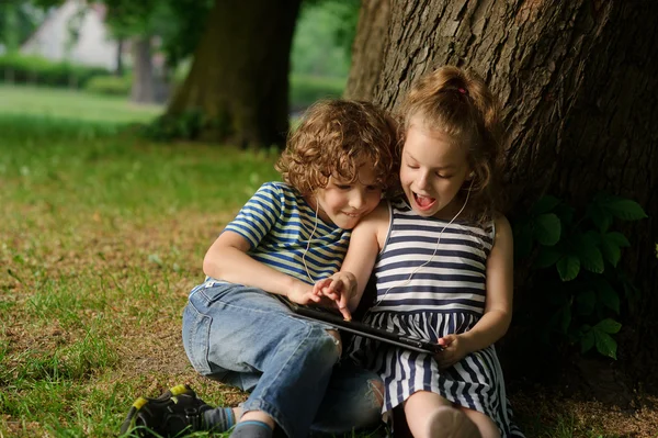 Broer met zus van 7-9 jaar zitten onder een boom en kijk in de laptop. — Stockfoto