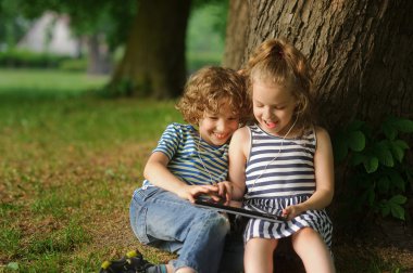 İki çocuk büyük bir ağaç altında parkta oturup tablet üzerinde oynamak.