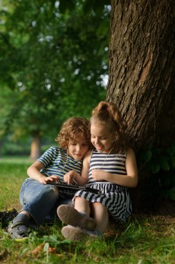 İki çocuk büyük bir ağaç altında parkta oturup tablet üzerinde oynamak.