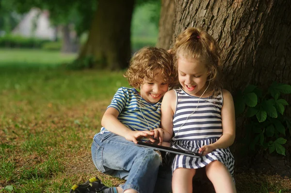 Twee kinderen zitten in park onder een grote boom en spelen op de Tablet PC. — Stockfoto