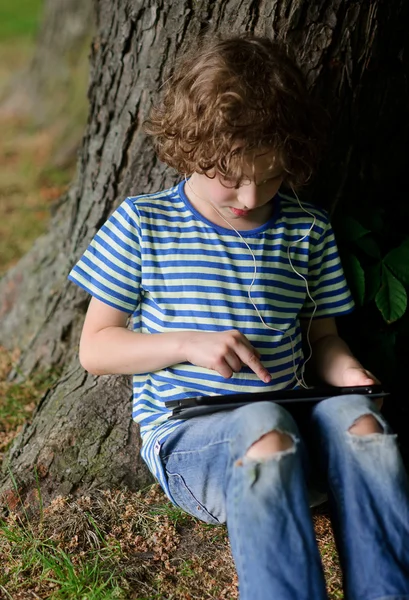 Chico se sienta apoyado en un tronco de árbol grande y viejo y mira a la pantalla de la tableta . — Foto de Stock