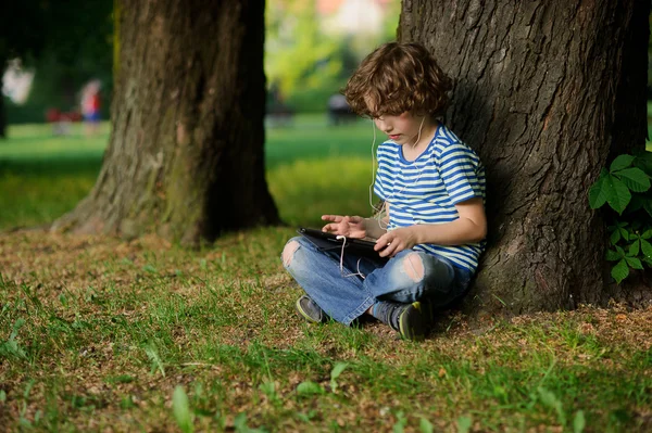 El niño de 8-9 años se sienta debajo de un gran árbol con la tableta en el regazo . — Foto de Stock