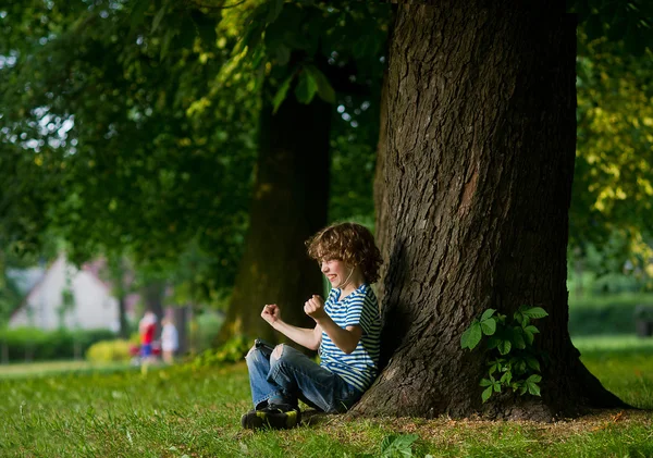 The boy with earphones in ears sits under a big tree.