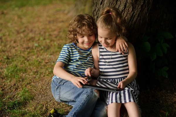 Twee kinderen zitten onder meer boom met interesse naar het tablet scherm. — Stockfoto