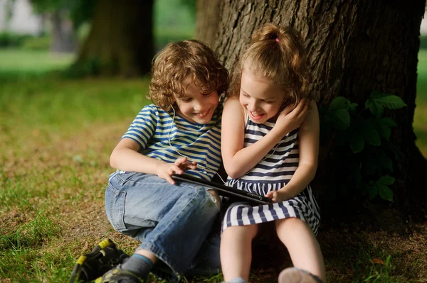 Niño con chica de 7-8 años sentarse bajo un árbol viejo y con entusiasmo mirar la pantalla del ordenador portátil . — Foto de Stock