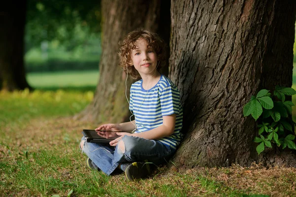 Мальчик 8-9 лет сидит, прислонившись к дереву и держит планшет в руке . — стоковое фото