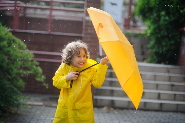 El niño divertido en un impermeable amarillo y con paraguas de pie en la lluvia . — Foto de Stock