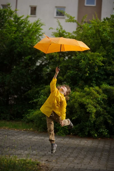 Boy in bright yellow raincoat flies over the earth with an umbrella in a hand. — Stock Photo, Image