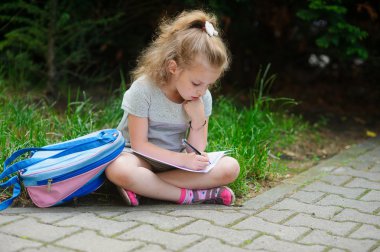 İlkokulun kız öğrenci bir çim üzerinde bacaklar çapraz olan oturur ve bir şey not defterine yazıyor.