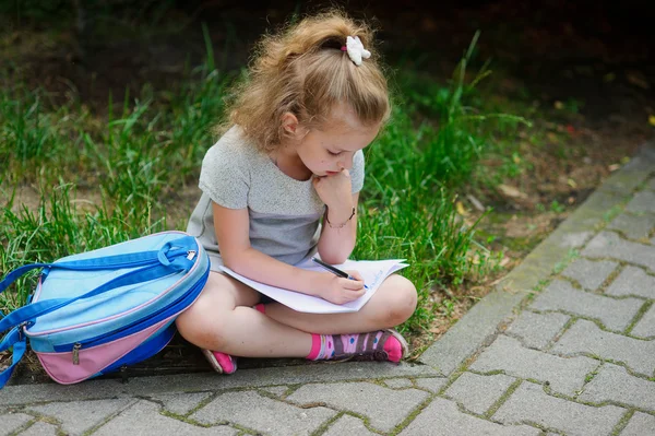 Das kleine Schulmädchen sitzt mit gekreuzten Beinen auf dem Gras und etwas schreibt in ein Notizbuch. — Stockfoto