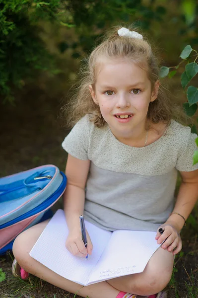 Kleines Schulmädchen sitzt mit gekreuzten Beinen unter einem Baum und hält auf dem Schoß ein offenes Notizbuch. — Stockfoto