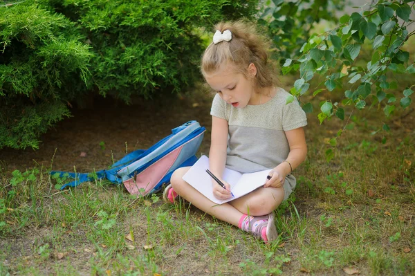 Erstklässler sitzt mit gekreuzten Beinen unter einem Baum und macht Hausaufgaben. — Stockfoto