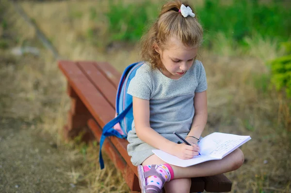 Маленькая школьная мисс сидит, скрестив ноги на скамейке в парке и что-то усердно пишет в блокноте . — стоковое фото