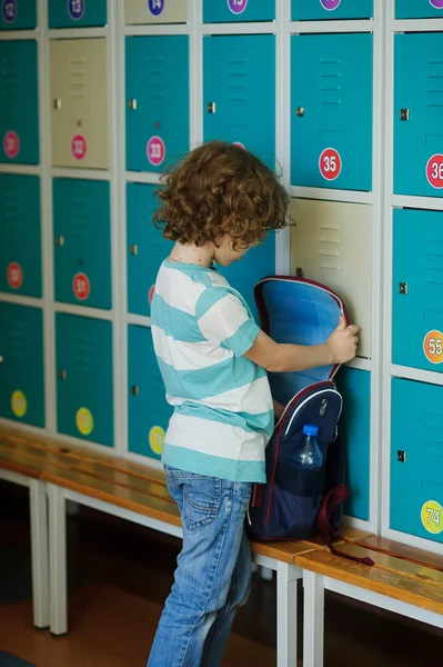 Little learner standing in the hall near the lockers and searching for something in his backpack. — Stock Photo, Image