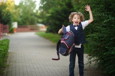 Cute little schoolboy, waving his briefcase in the schoolyard. clipart