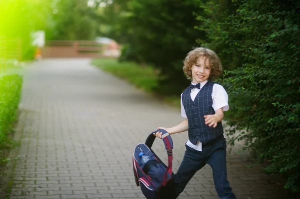 Carino piccolo scolaro, agitando la valigetta nel cortile della scuola . — Foto Stock