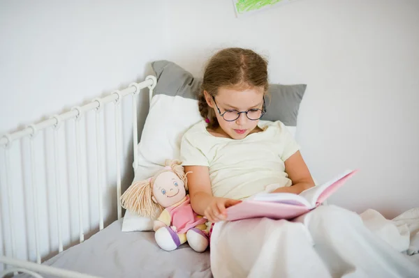 Little girl in glasses reading a book while lying in bed. — Stock Photo, Image