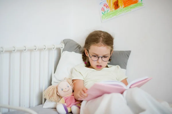 Niña en gafas leyendo un libro mientras está acostada en la cama . —  Fotos de Stock