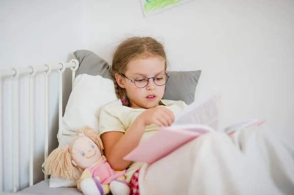 Niña con gafas hojeando un libro mientras está acostada en la cama . —  Fotos de Stock