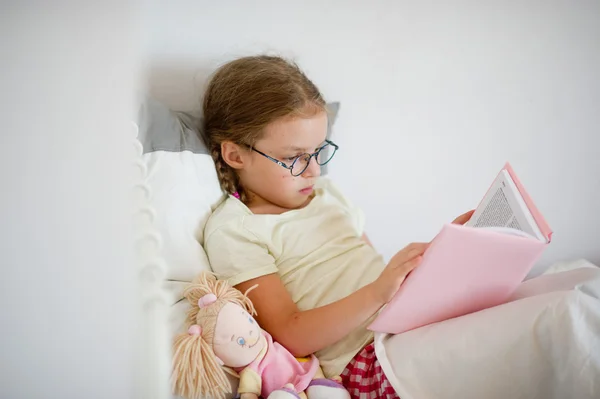 Little girl with pigtails reading a book while lying in bed. — Stock Photo, Image