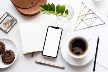 Akıllı telefon modeli, kahve fincanı ve not defteri olan beyaz ofis masası.