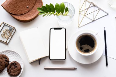 Akıllı telefon modeli, kahve fincanı ve not defteri olan beyaz ofis masası.