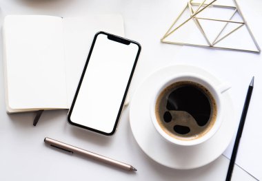 Akıllı telefon modeli, kahve fincanı ve not defteri olan beyaz ofis masası.