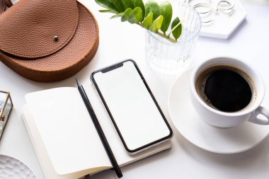 Akıllı telefon modeli, kahve fincanı ve not defteri olan beyaz ofis masası.