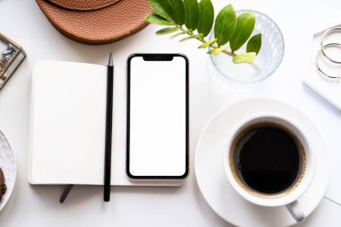 Akıllı telefon modeli, kahve fincanı ve not defteri olan beyaz ofis masası.