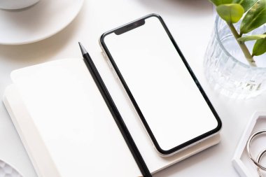 Akıllı telefon modeli, kahve fincanı ve not defteri olan beyaz ofis masası.