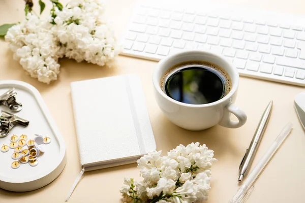 Beige and white work space with coffee cup, notepad and lilac flowers — Stockfoto