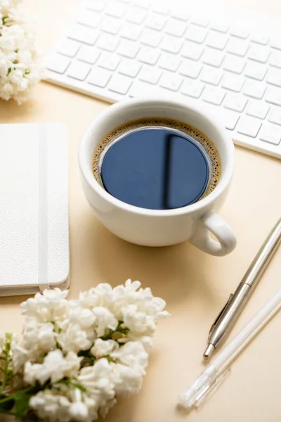 Beige and white work space with coffee cup, notepad and lilac flowers