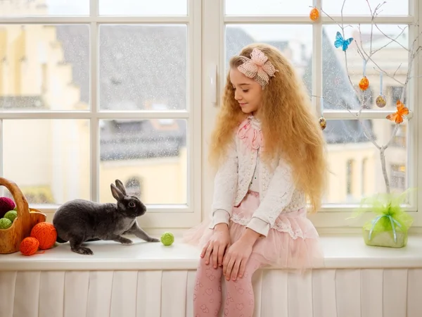 Hermosa niña sentada en un alféizar de la ventana y jugando con el conejo de Pascua . —  Fotos de Stock