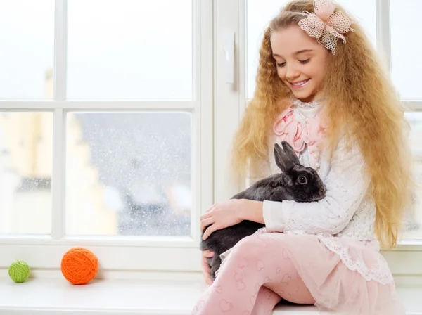 Hermosa niña sentada en un alféizar de la ventana y jugando con el conejo de Pascua . — Foto de Stock