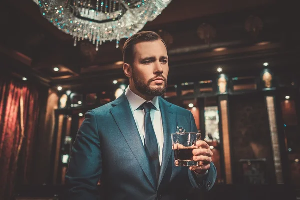 Hombre seguro y bien vestido con un vaso de whisky en el interior de un apartamento de lujo . — Foto de Stock