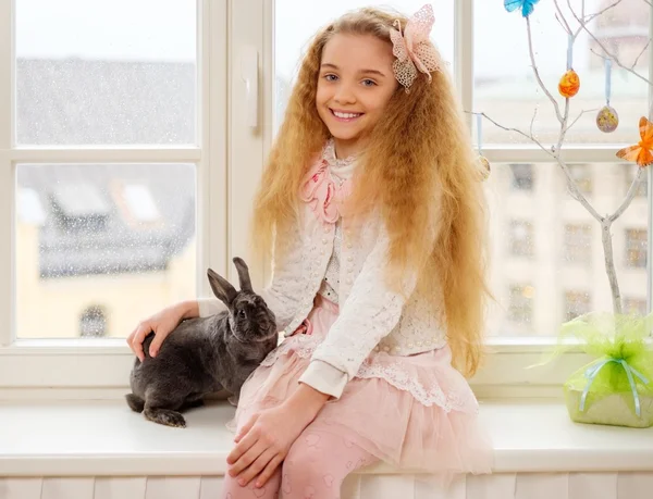 Hermosa niña sentada en un alféizar de la ventana y jugando con el conejo de Pascua . —  Fotos de Stock