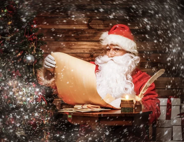 Santa Claus in wooden home interior reading wish list scroll — Stock Photo, Image